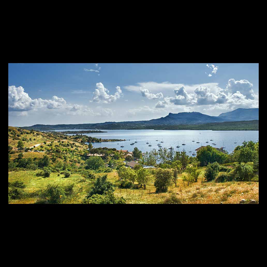 El tranquilo embalse del Atazar resguarda los barcos de recreo en su puerto bajo un cielo azul cubierto de grandes nubes blancas