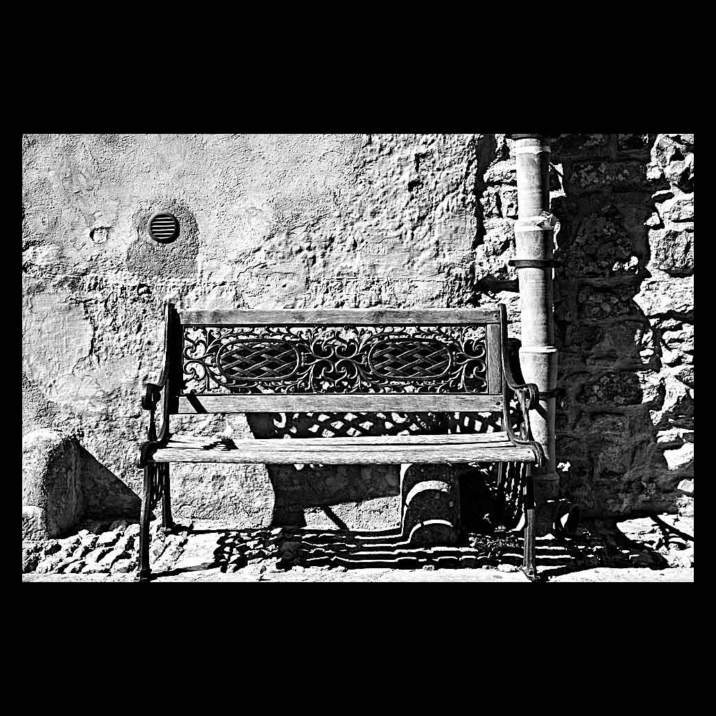 Banco vacío de madera y hierro forjado en una calle de Sicilia. Fotografía en blanco y negro con sombras marcadas y texturas de pared de piedra antigua.