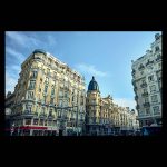 Fachadas de edificios clásicos en la Gran Vía de Madrid bajo un cielo azul despejado. Destaca la arquitectura ecléctica, los balcones de piedra y una cúpula negra al fondo en la zona de la Red de San Luis.