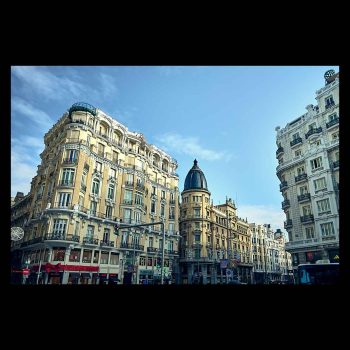 Fachadas de edificios clásicos en la Gran Vía de Madrid bajo un cielo azul despejado. Destaca la arquitectura ecléctica, los balcones de piedra y una cúpula negra al fondo en la zona de la Red de San Luis.