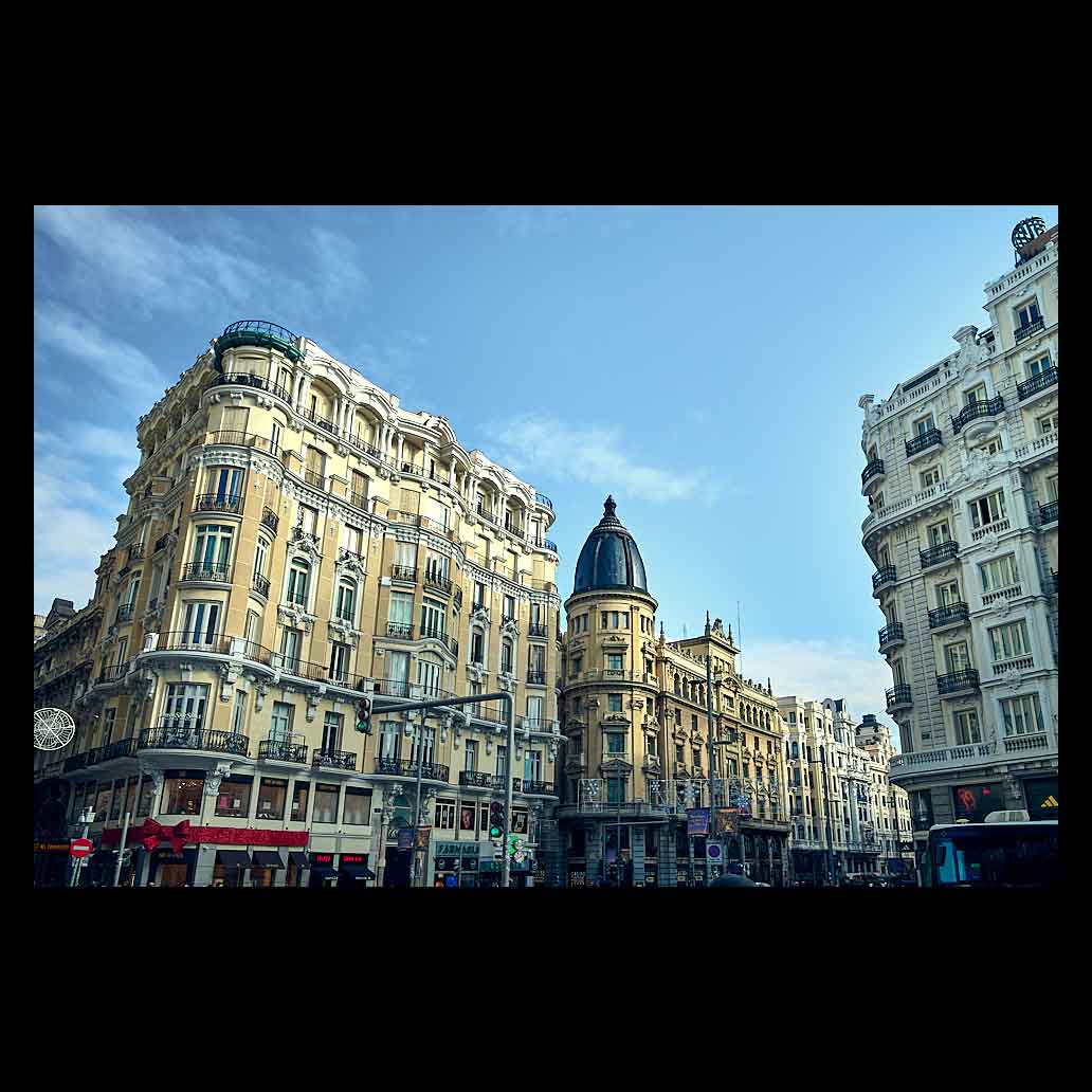 Fachadas de edificios clásicos en la Gran Vía de Madrid bajo un cielo azul despejado. Destaca la arquitectura ecléctica, los balcones de piedra y una cúpula negra al fondo en la zona de la Red de San Luis.