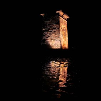 Fotografía nocturna del Templo de Debod en Madrid iluminado en tonos cálidos, reflejándose sobre el agua oscura con ondas suaves.